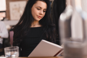 Young beautiful brunette woman reading interesting book in cafe. Girl smiling and look joyful. Student teen girl studying in restaurant.