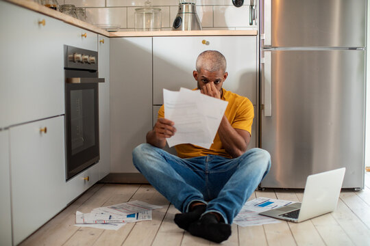 Stressed man sitting on kitchen floor with bills and laptop