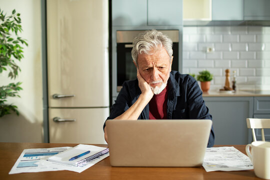 Worried senior man using laptop with bills on desk at home