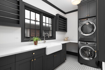 A laundry room with dark grey cabinets, grey over under washer and dryer, farmhouse sink, pattern...