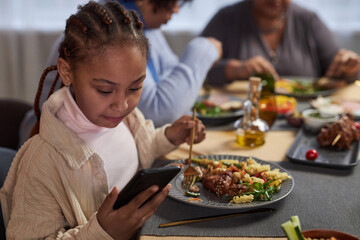 Portrait of little Black girl using smartphone at dinner table while eating at home with family 