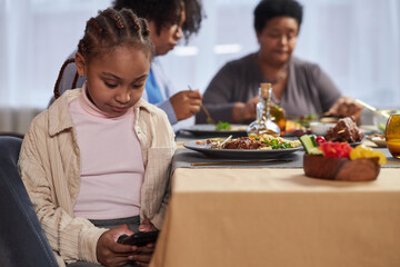 Portrait of young Black girl using smartphone under table at dinner and typing text messages copy...