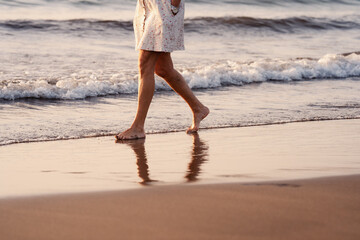 An elderly woman in a dress walks alone on the beach at sunset, reflecting on the wet sand. Senior...