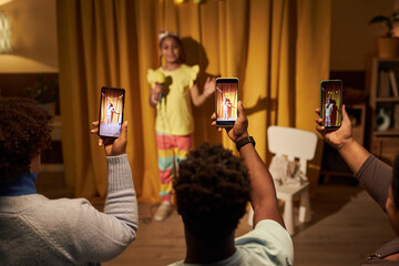 Black little girl performing in home concert or talent show with parents holding smartphone and...