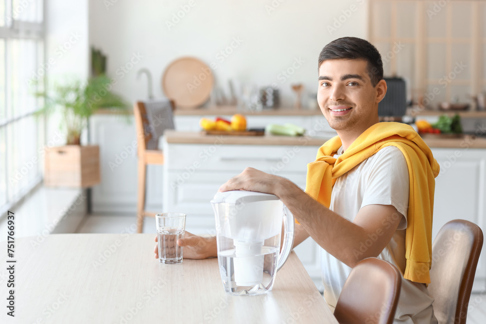 Wall mural young man with filter jug and glass of fresh water sitting at table in kitchen