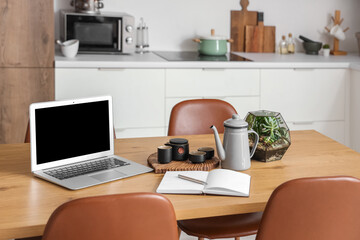 Laptop with blank screen, tea set and notebook on table in modern kitchen