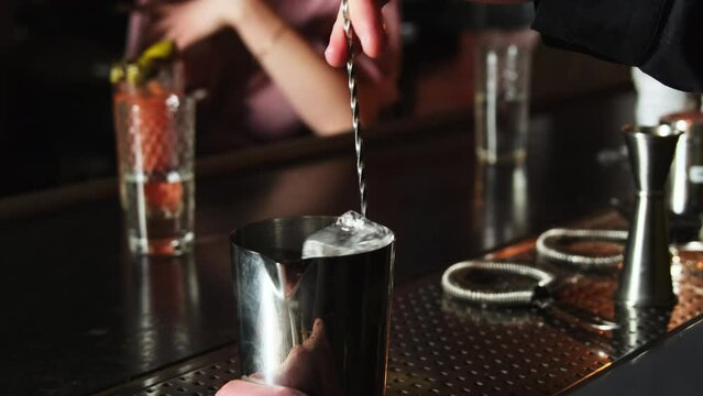 Barkeeper stirs ice cubes in aluminum glass with long bar spoon. Cocktail preparation process on bar counter against customers