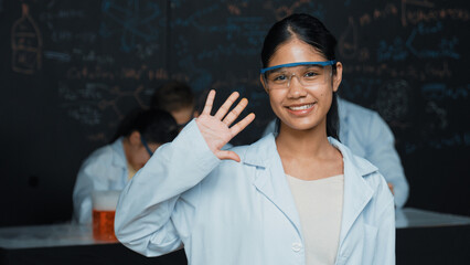 Cute girl waving hands to camera while people doing experiment at laboratory. Young cute student...
