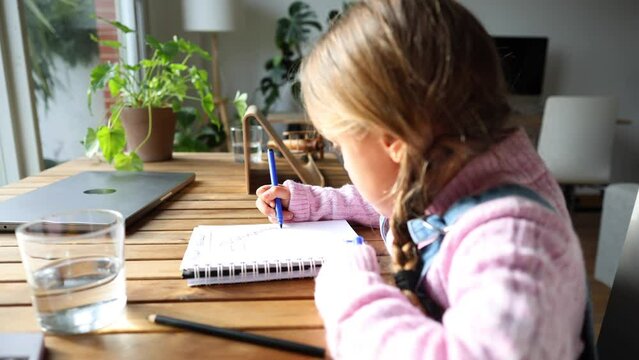 A smart little Caucasian girl sitting at the table at home paints in a notebook near the window. Learning, education concept.