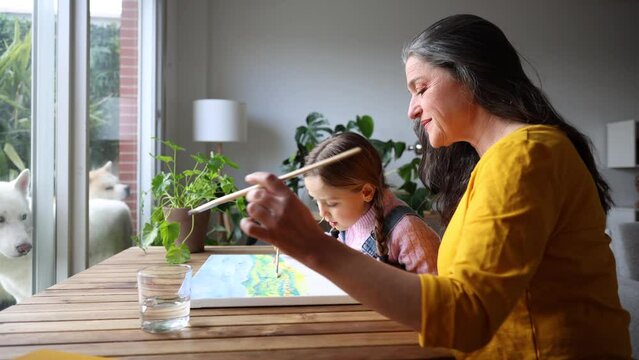 grandmother and granddaughter painting a picture together in the living room in front of the window
