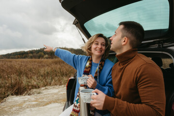 Happy young woman and man sitting in the open trunk of a car while traveling in autumn, road trip concept