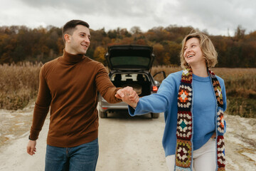 Smiling couple holding hands, strolling down country road with car on the background, happy man and woman on a roadtrip