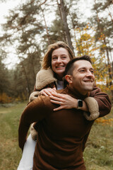 Adult couple having fun in the autumn park outdoors, happy man is carrying a woman on his back in the woods