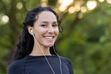 Optimistic diverse sportswoman in black top smiling at camera while listening to motivating playlist in wired earphones. Attractive female in black top finishing training in green public area.
