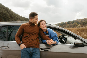 Happy man leaning to a car and holding hands of cheerful woman that sitting inside, adult couple enjoying road trip in autumn