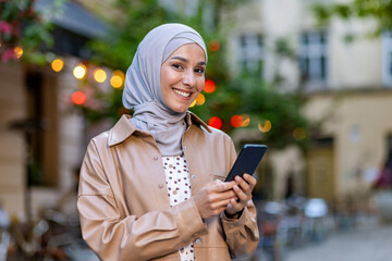Adult arab woman wearing pastel hijab and holding modern smartphone while having walk on central...