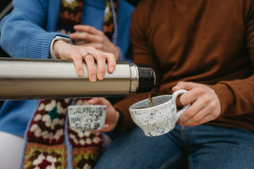 A man and woman drinking tea from thermos during autumn travel, couple holding handmade cups filled with warm drink