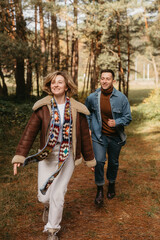 Happy couple is having fun in the middle of the forest, a smiling woman is running with her husband in an autumn park.