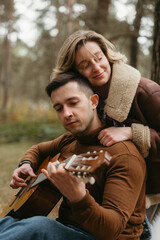 Happy couple spending time together in autumn park, man playing guitar while woman hugs him