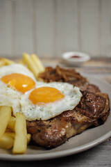 Bistec a lo pobre. Beef, eggs and fried potatoes on a plate on a wooden table with cutlery. Vertical close-up. Typical Chilean food