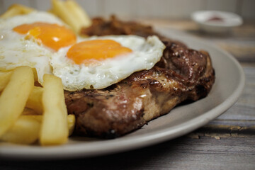 Bistec a lo pobre. Beef, eggs and fried potatoes on a plate on a wooden table with cutlery. Close-up. Typical Chilean food