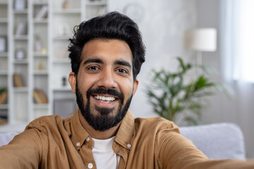 Portrait of indian male with stylish hairstyle taking selfie and holding camera with hands in light...