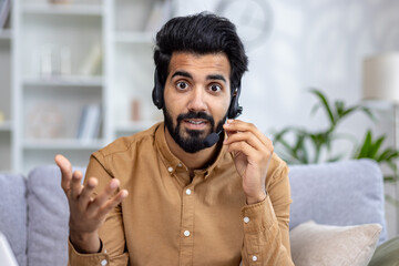 Enthusiastic latino male wearing professional headset with microphone and gesturing to camera during video call. Confident remote worker explaining information while fixing audio garniture indoors.