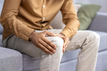 Close up of unknown person holding knee with both hands while sitting at grey couch with cushions. Adult man with leg trauma suffering from joint pain and massaging sore spot for facilitation.