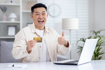 Cheerful asian man making like sign with hand while sitting at domestic office with laptop and...