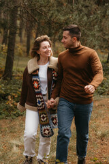 A happy man and woman are walking through the autumn forest, holding hands, adult couple spending time together outdoors