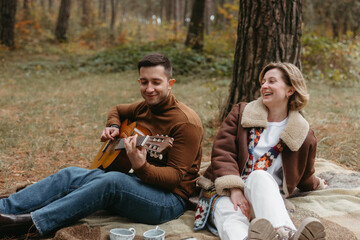 A man is playing a guitar for a happy woman while they are sitting on a blanket in the autumn woods