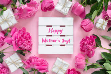 Bouquet of beautiful pink peonies with gift boxes in paper wrapping. Round frame of flowers. White wooden board with text Happy Mother's Day!