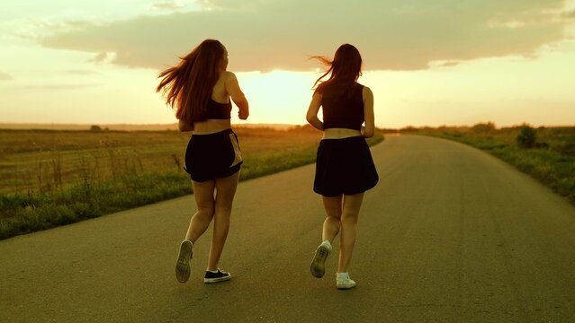 Athletic young women running along an asphalt road at sunset, healthy fitness lifestyle. Girls run along road, training teamwork of running athletes. Slow motion. People run in sun together. Sport run