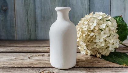 Rolgordijnen a white ceramic bottle with a dry hydrangea flower stands on a wooden surface © Deanne