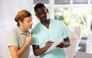 Experienced African American male doctor consulting interested man patient filling medical form before consultation in clinic