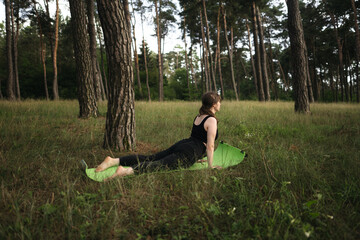 Yoga cobra pose by woman in black suit on green grass in the park around pine trees. Alone yoga in the garden in the morning, stretch back. Healthy lifestyle. 