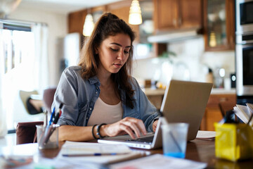 Focused young woman managing finances and paying bills on her laptop at the kitchen table, surrounded by paperwork.

