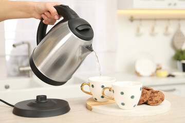 Woman pouring hot water from electric kettle into cup in kitchen, closeup. Space for text