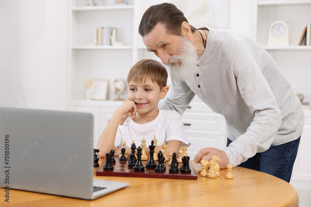 Poster Grandfather teaching his grandson to play chess following online lesson at home