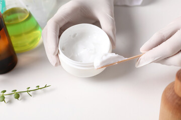 Dermatologist with jar testing cosmetic product at white table, closeup