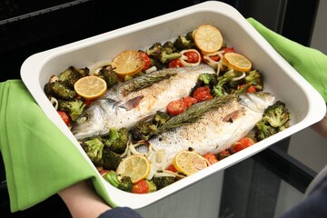 Woman taking baking dish with delicious fish and vegetables from oven, closeup