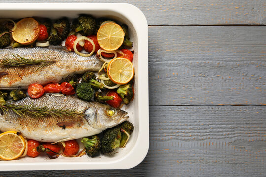 Delicious fish with vegetables and lemon in baking dish on grey wooden table, top view. Space for text