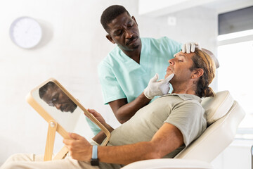 Experienced African male cosmetologist examines facial skin of man patient and advises on...