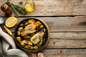 Pieces of delicious baked cod with vegetables, lemon and spices in dish on wooden table, flat lay. Space for text