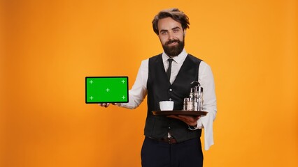 Joyful employee shows greenscreen on tablet, posing with food platter against yellow background....