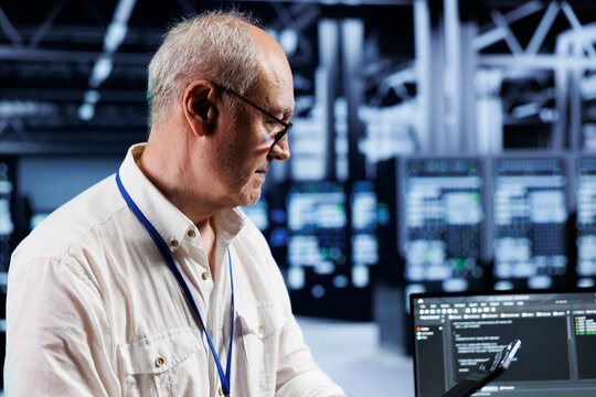 Old technician coding in technologically advanced data center using javascript language. Skilled specialist updating software on server cabinets using laptop, ensuring optimal performance and security