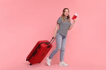 Happy young woman with passport, ticket and suitcase on pink background