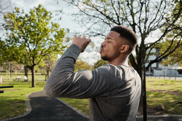 Fototapeta premium Young black man in sports clothing with closed eyes drinking water from bottle in park