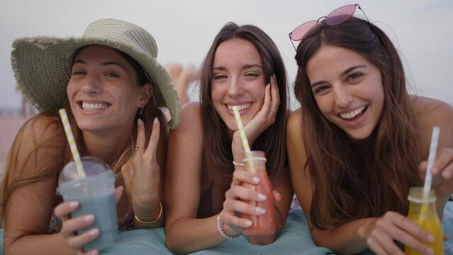 portrait of three caucasian girls looking at camera and smiling. They drink natural smoothies. These attractive women look carefree and enjoy their vacation during the summer posing outdoors
