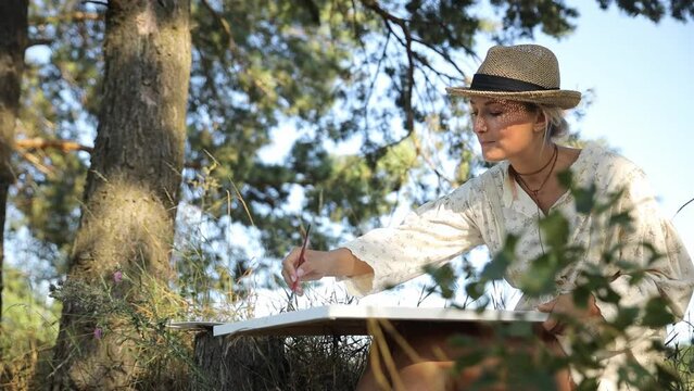 A woman in a hat is painting a picture of a tree